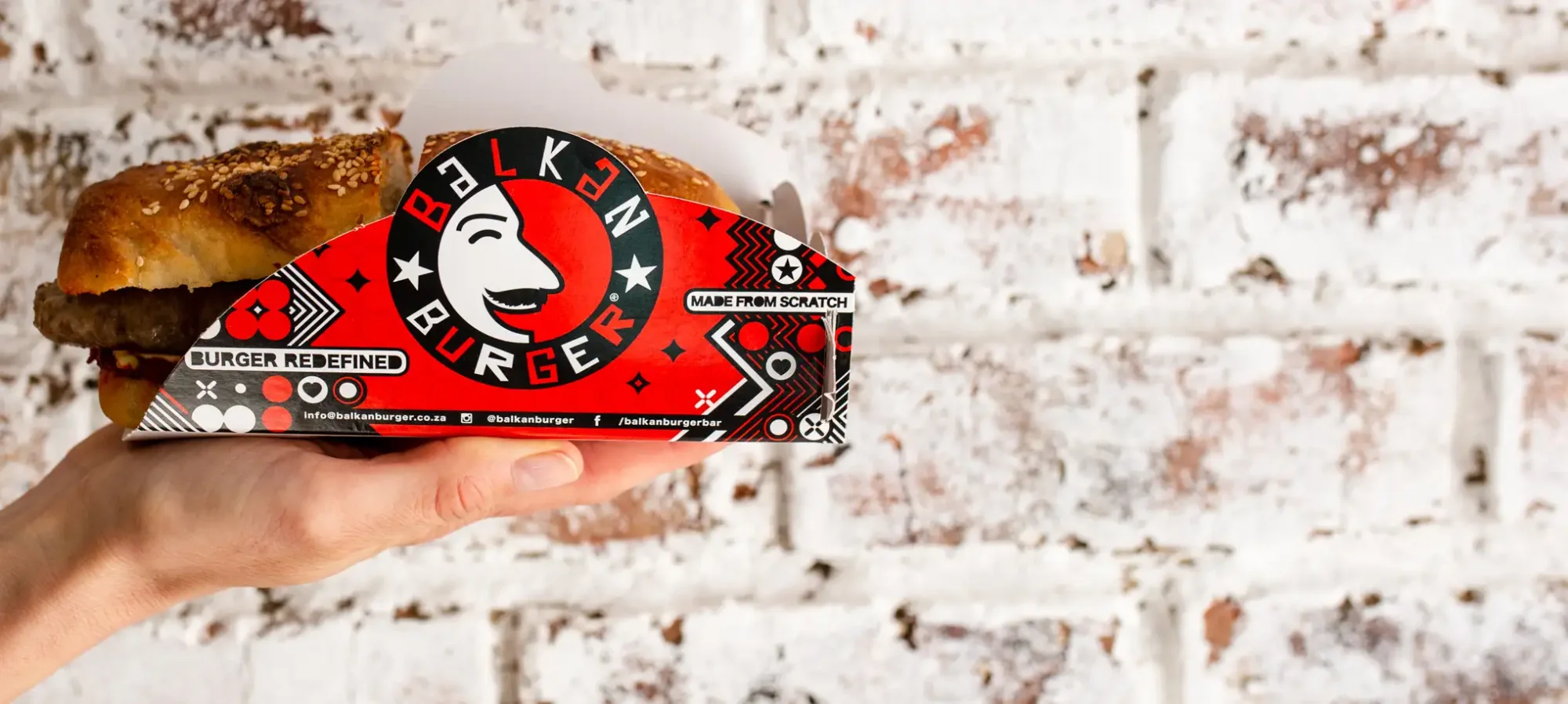 Hand holding a red Balkan Burger box against a brick wall.