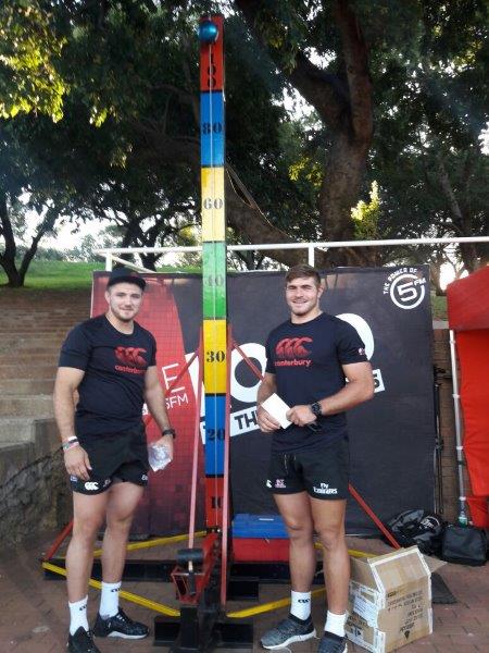 Two men in black shirts next to a height measurement tower outdoors.