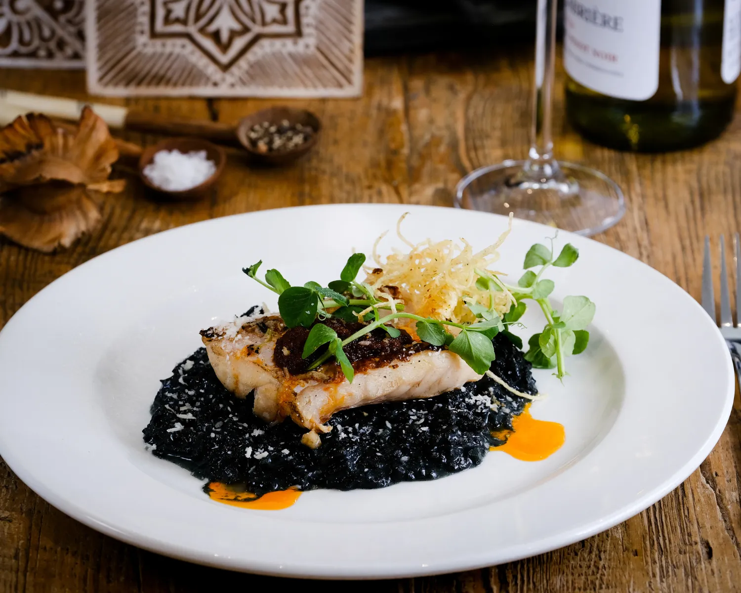 Plated fish fillet on black risotto with garnishes.