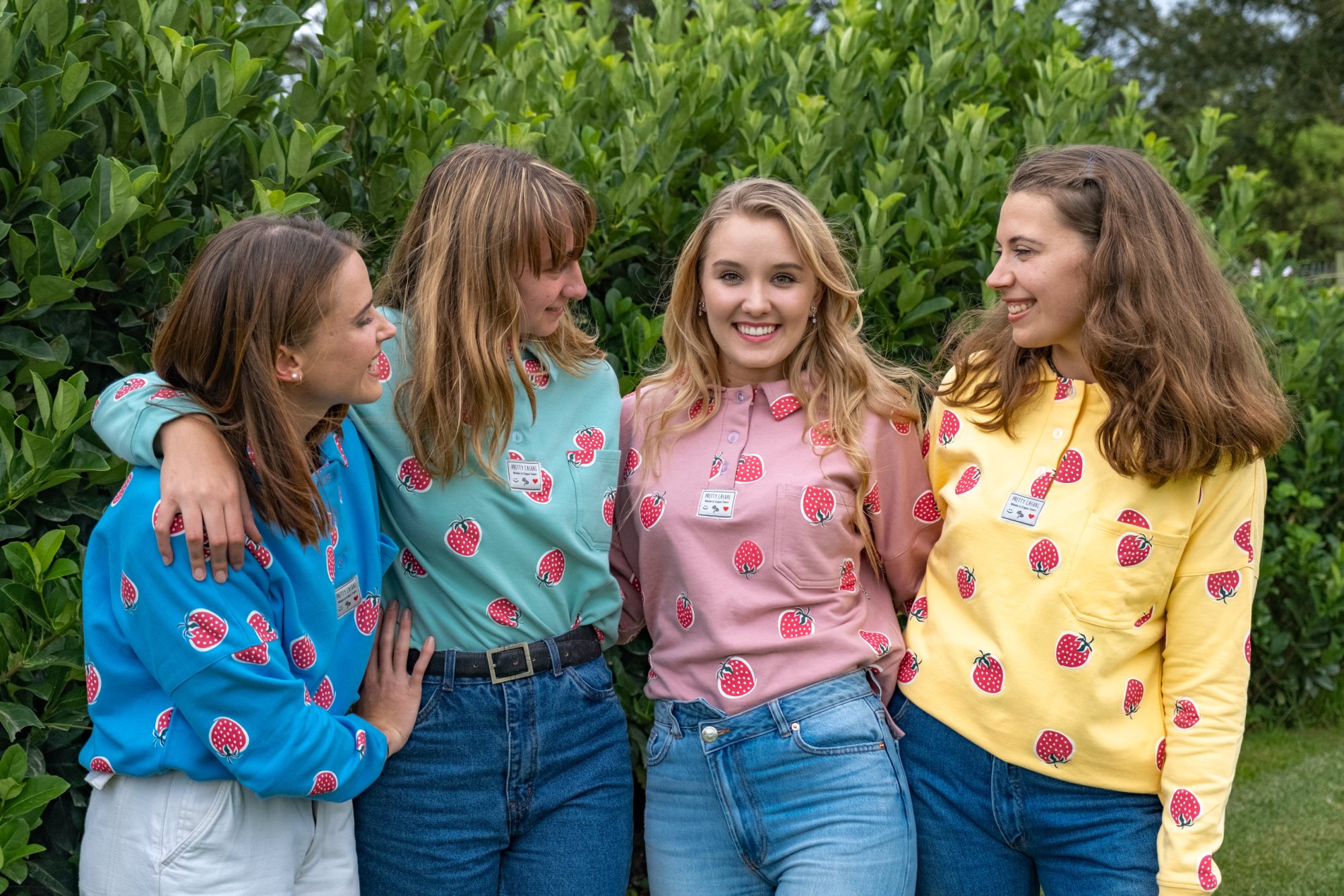 Four young women wearing strawberry-patterned sweatshirts in front of green foliage