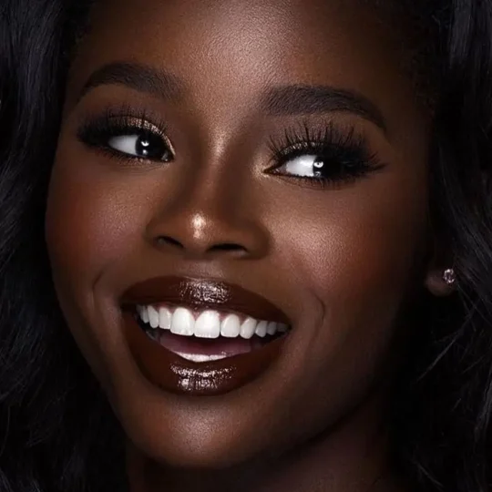 Close-up of a woman smiling with glam makeup, featuring dark lips and long lashes.
