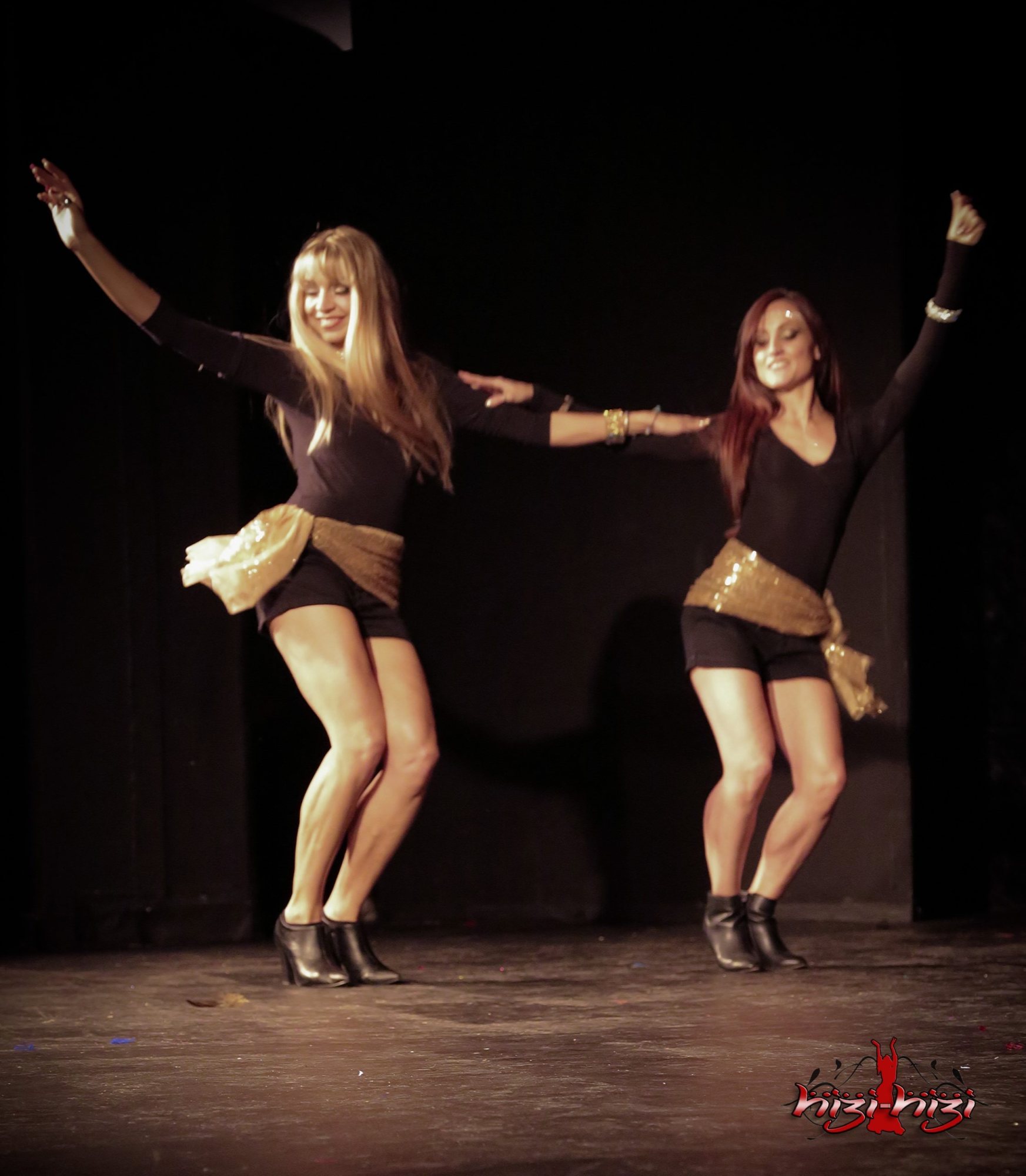 Two belly dancers in black outfits performing energetically on stage.