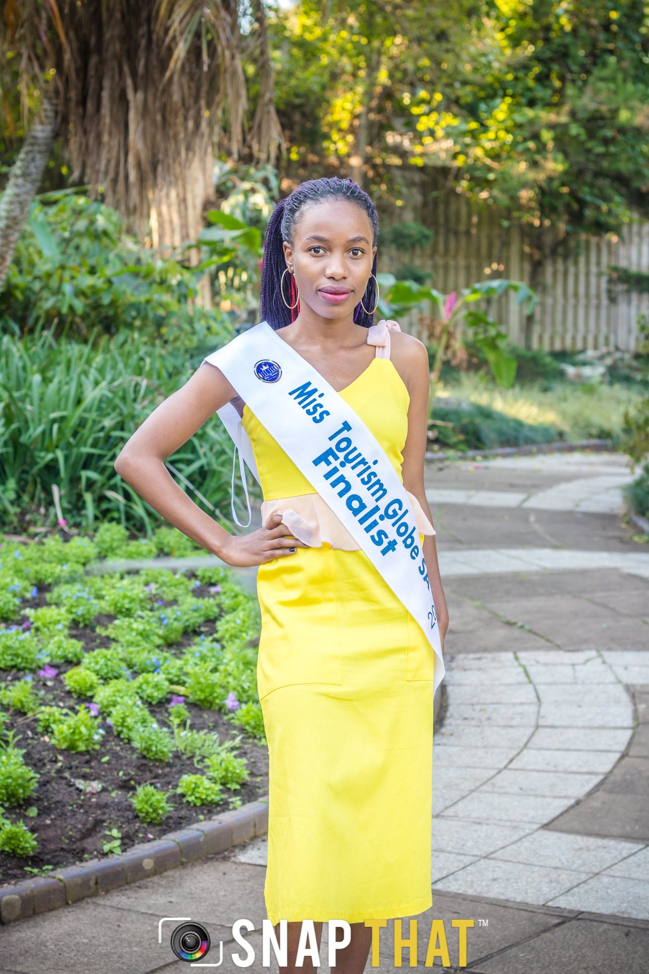 A contestant in a yellow dress with a sash standing in a garden