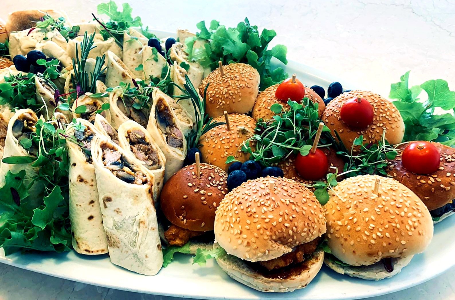 A platter with wraps and mini burgers garnished with greens and tomatoes