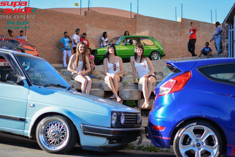 Three women seated among cars at a vibrant car show.