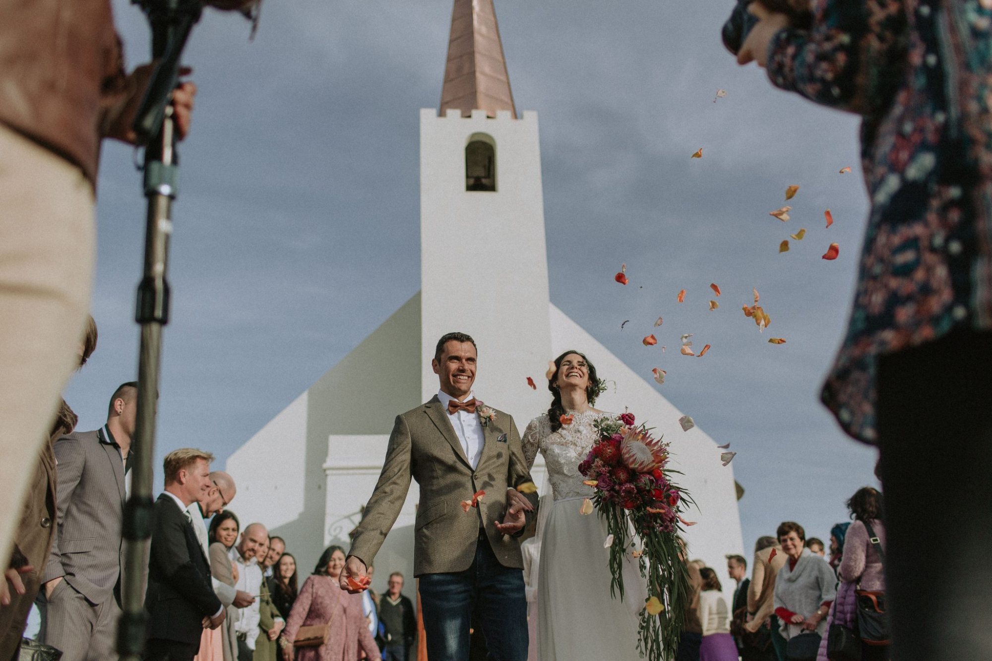 Couple celebrates wedding outside a white chapel with flower petals in the air.