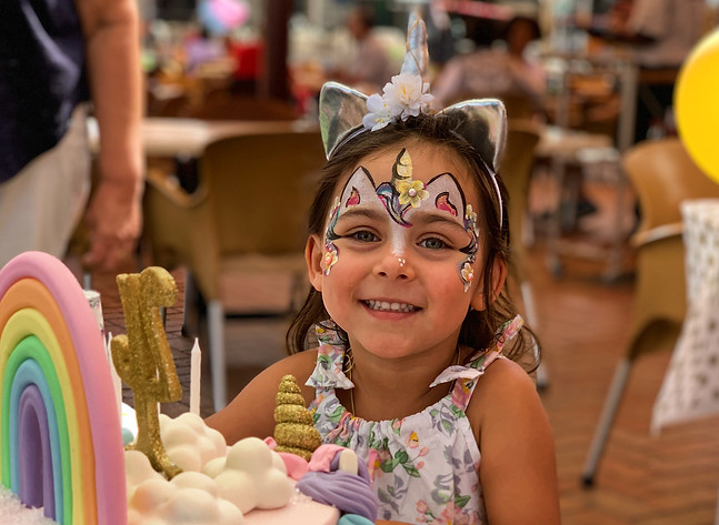 Child with face painted as unicorn beside a colorful cake