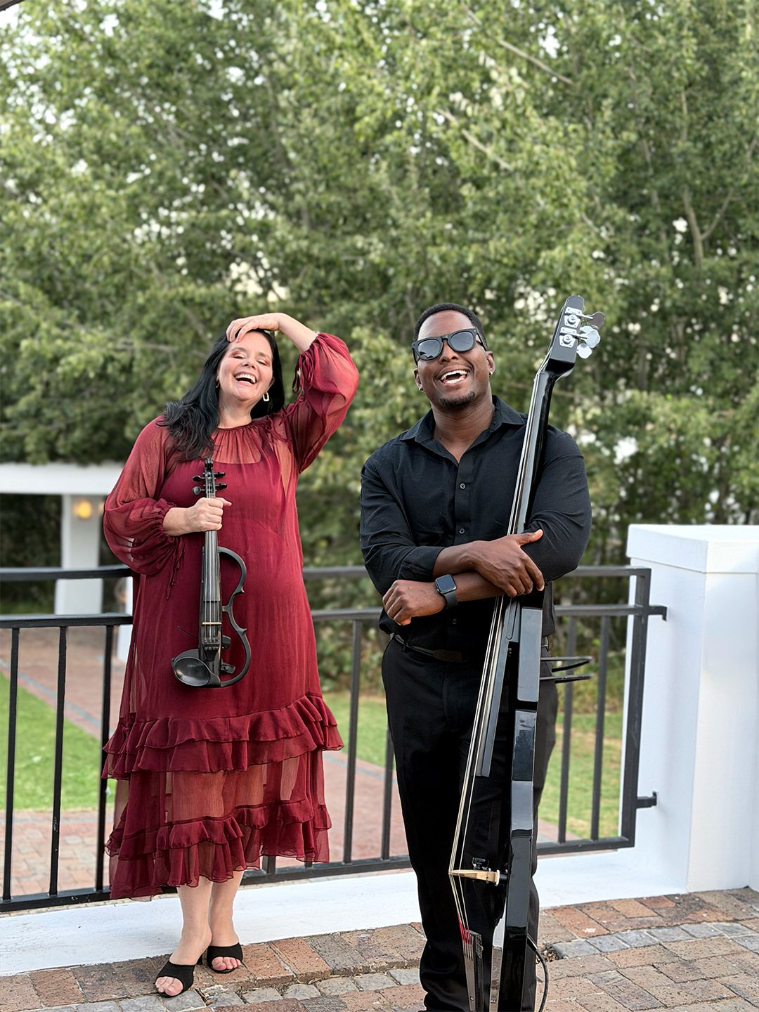 Two musicians, a female violinist and a male bassist, smiling outdoors.