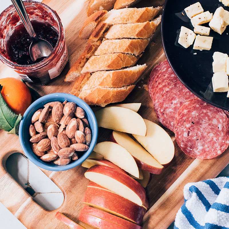 A platter with bread, salami, apple slices, almonds, cheese cubes, and jam.