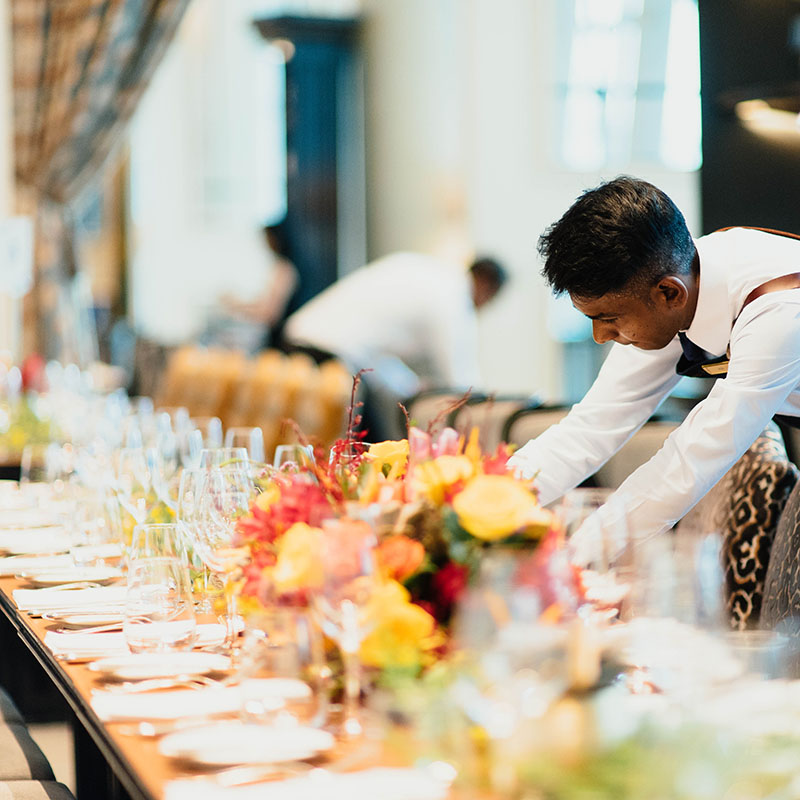 Waiter arranging an elegant dining table with flowers and glassware.