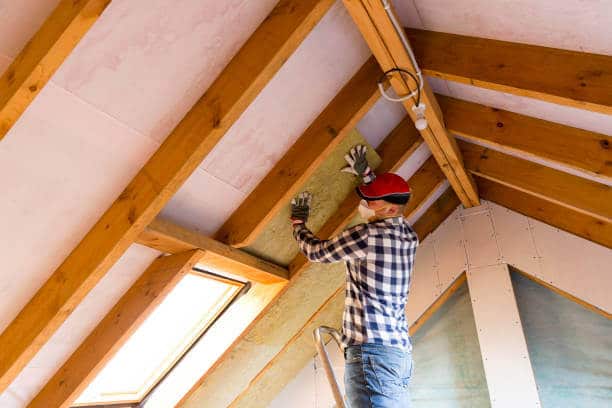 A worker installing insulation in an attic with wooden beams.